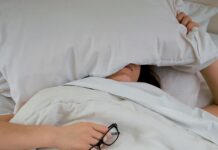 피로회복을 위한 에너지 충전 person lying on bed while covering face with pillow and holding eyeglasses